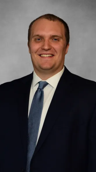 Dr. Andrew Arnold poses for professional headshot wearing a black suit and blue tie in front of a gray background.