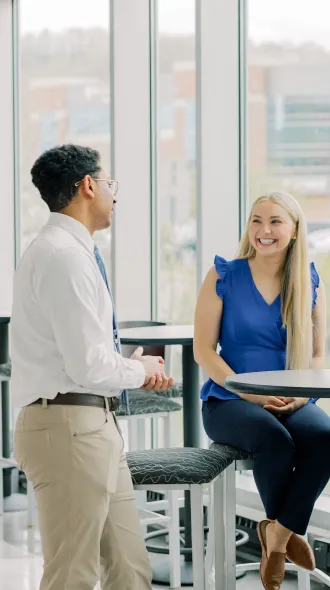 Four people in business attire are engaged in a lively discussion around a table by large windows, with a modern building visible in the background.
