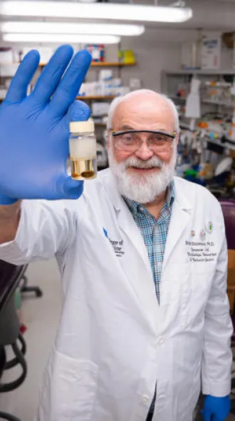 Brian Stevenson wearing white coat, safety glasses, and gloves. He stands in his lab, smiling, and holding up a sample to the camera.
