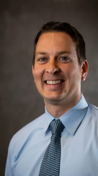 Dr. Lance Johnson headshot (A smiling man in a light blue dress shirt and patterned tie poses against a dark, neutral background.)