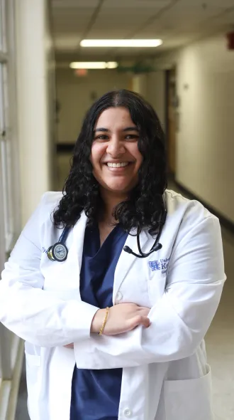 Supriya Challa stands with arms crossed in hallway, smiling, wearing a white coat