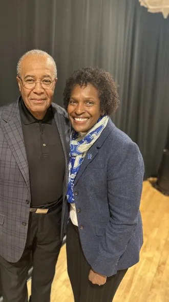 Dr. Lovoria Williams and Pastor Joseph Owen pose together, smiling warmly. Dr. Williams wears a navy blazer with a blue and white scarf, while Pastor Owen wears glasses, a dark polo shirt, and a gray plaid blazer. They stand in front of a black curtain in an indoor venue.