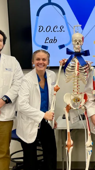 Five people in lab coats and blue attire stand smiling beside a skeleton model wearing a bow tie. A sign behind reads "D.O.C.S. Lab."