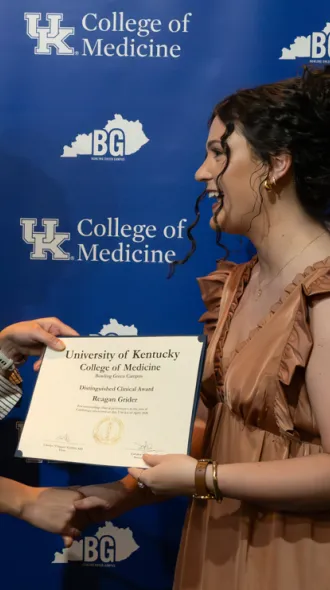 Two smiling individuals exchange a certificate in front of a University of Kentucky College of Medicine backdrop, conveying achievement and celebration.