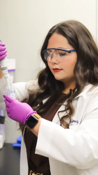 A scientist in a lab coat uses a pipette in a lab, wearing safety goggles and purple gloves. Shelves with lab equipment and microscopes are around her.