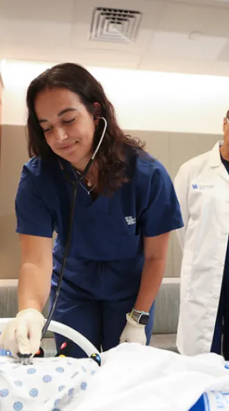 Medical simulation with two med students in blue scrubs and a white coat attending to a patient mannequin in a hospital room setting.