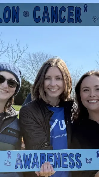 In a large cardboard frame that reads "Colon Cancer Awareness" three women smile. One holds a baby.