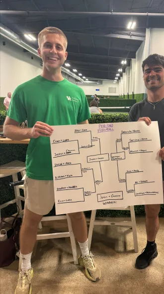 Two men smiling and holding a tournament bracket poster titled "Paso Fino Pickleball." They're in an indoor sports venue. 