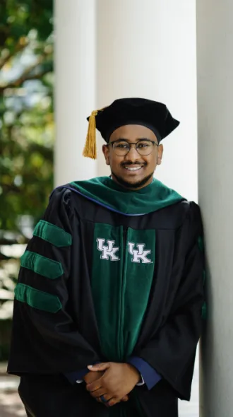 Kidus stands smiling in academic regalia with green accents and a gold tassel, leaning against a column