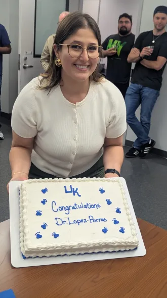 A brunette woman holds a white cake with blue paw prints and UK, it says "Congratulations Dr. Lopez-Porras"