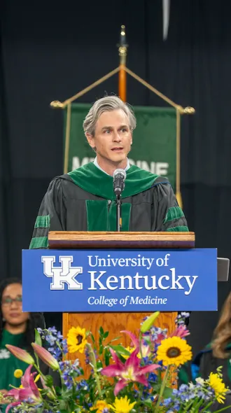 Brent Morris, MD, wears regalia and stands behind a UK College of Medicine-labeled podium at graduation