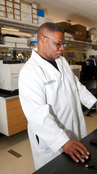 Daniel Lee, wearing a white lab coat, works at a computer beside research equipment in a laboratory.