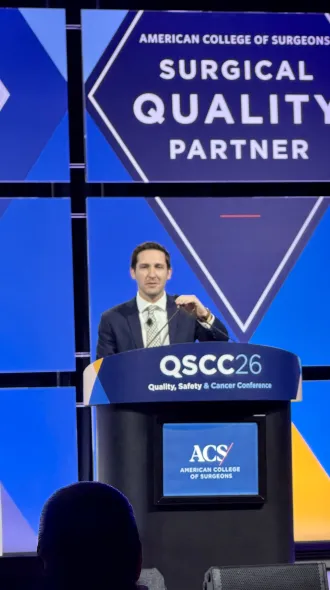 Dr. Jacob Hubbuch, in blue blazer and tie, stands behind podium labeled QSCC (stands for Quality, Safety, and Cancer Conference) while projected behind him is a blue diamond reading "American College of Surgeons Surgical Quality Partner."