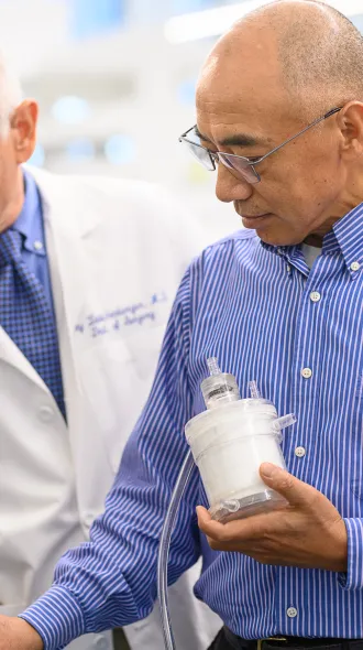 Dr. Joseph Zwischenberger, an older doctor dressed in white lab coat, looks at a clear box with wires and electronic displays. Next to him is  Dr. Dongfang Wang in a blue button down shirt demonstrates the function of a white medical assist device he holds in his hand that is wired to the box