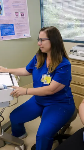 A health care professional in blue scrubs uses a computer beside a seated pregnant patient in an exam room.