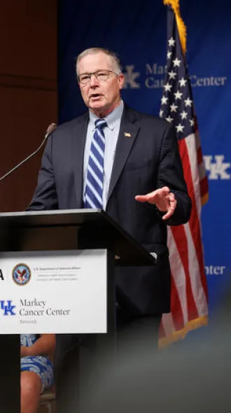 A person wearing glasses, a dark suit and striped blue tie speaks at a podium during a formal event. An American flag and a blue backdrop displaying the UK Markey Cancer Center logo are behind the speaker.