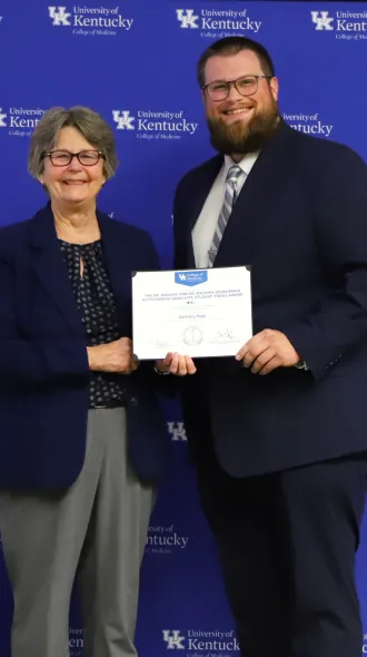 Dr. Garvy and Zachary Kipp pose in front of a UK College of Medicine backdrop, smiling. He holds a certificate. 